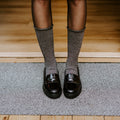 Cropped view of slender legs in Solid Bamboo socks in Static paired with glossy black loafers, standing on a textured doormat at the threshold of a sunlit wood-floored entryway. [lf]