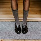 Cropped view of slender legs in Solid Bamboo socks in Static paired with glossy black loafers, standing on a textured doormat at the threshold of a sunlit wood-floored entryway. [lf]