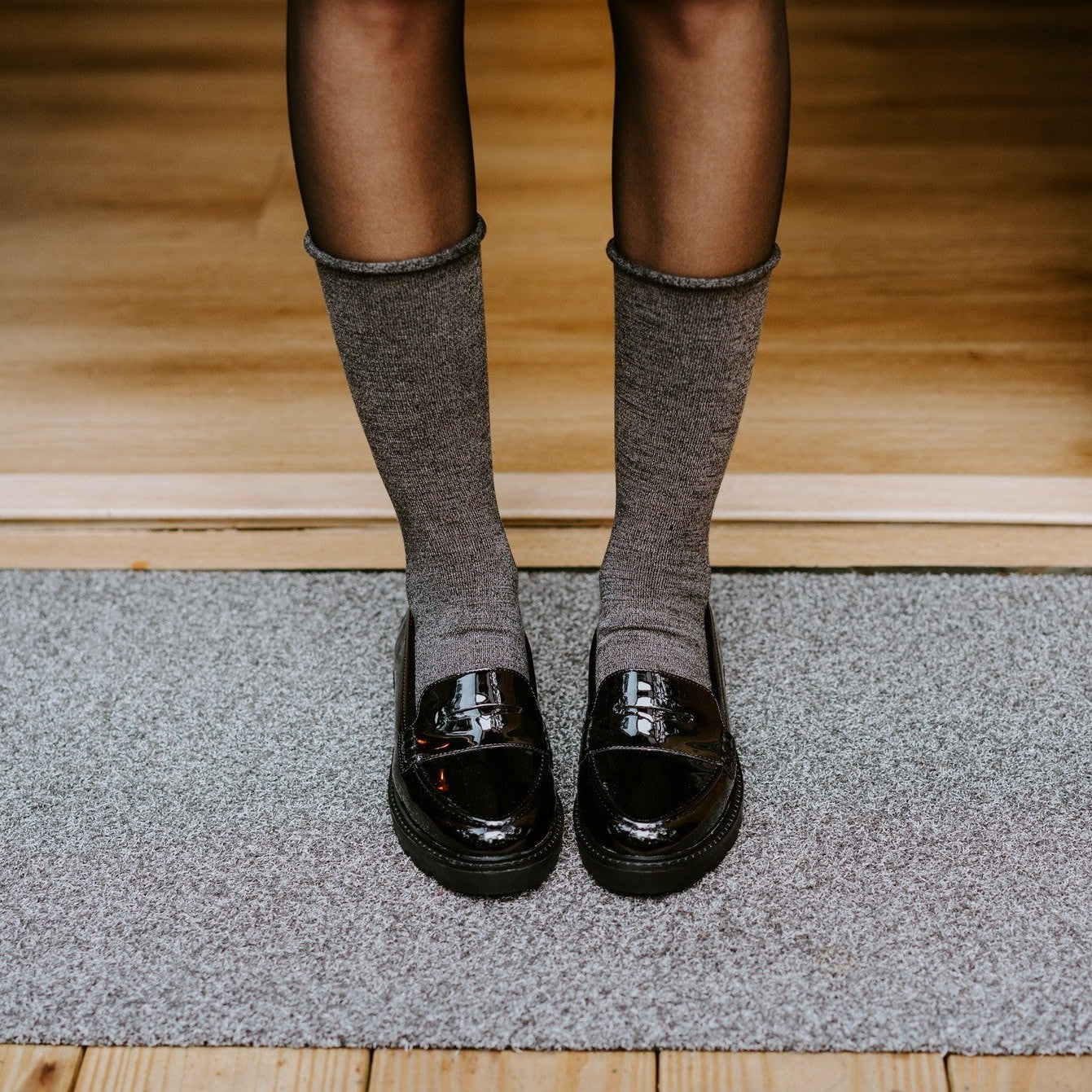 Cropped view of slender legs in Solid Bamboo socks in Static paired with glossy black loafers, standing on a textured doormat at the threshold of a sunlit wood-floored entryway. [lf]