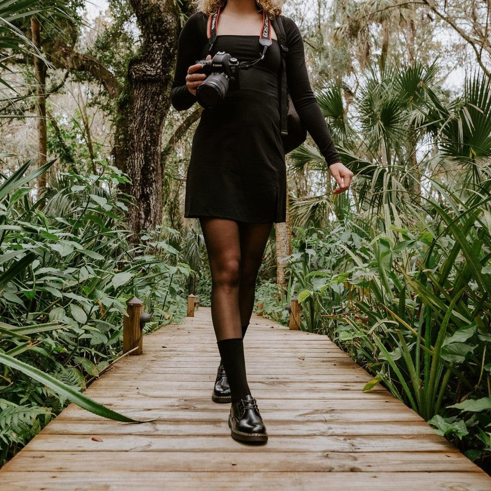 Smiling woman in a long-sleeve black dress walks straight toward the camera holding her DSLR, Solid Bamboo socks in Static rising above polished black boots on the boardwalk path. [lf]