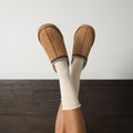 Legs raised against a white wall in chestnut suede slippers with Solid Bamboo socks in Ivory peeking above the cuffs over dark wood flooring. [lf]