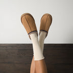 Legs raised against a white wall in chestnut suede slippers with Solid Bamboo socks in Ivory peeking above the cuffs over dark wood flooring. [lf]