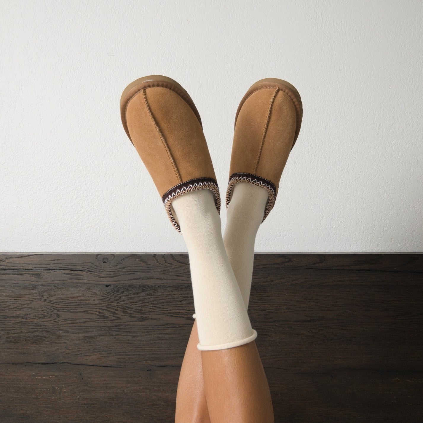 Legs raised against a white wall in chestnut suede slippers with Solid Bamboo socks in Ivory peeking above the cuffs over dark wood flooring. [lf]