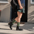 A vertical full body frame of the same model captured mid stride in front of the dark door, the Solid Bamboo socks in Green Heather visible between her plaid skirt and black lug sole Chelsea boots. [lf]