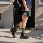 A vertical full body frame of the same model captured mid stride in front of the dark door, the Solid Bamboo socks in Green Heather visible between her plaid skirt and black lug sole Chelsea boots. [lf]