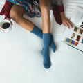 A model sits cross legged on a white bed holding a black mug of coffee beside an open art reference book, her crossed ankles wearing Solid Bamboo socks in Blue. [lf]