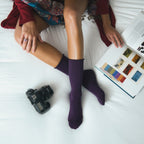 A relaxed bedroom scene with a paisley shawl draped over a red top, a vintage camera beside the model's legs, and Solid Bamboo socks in Purple worn above pointed toes. [lf]