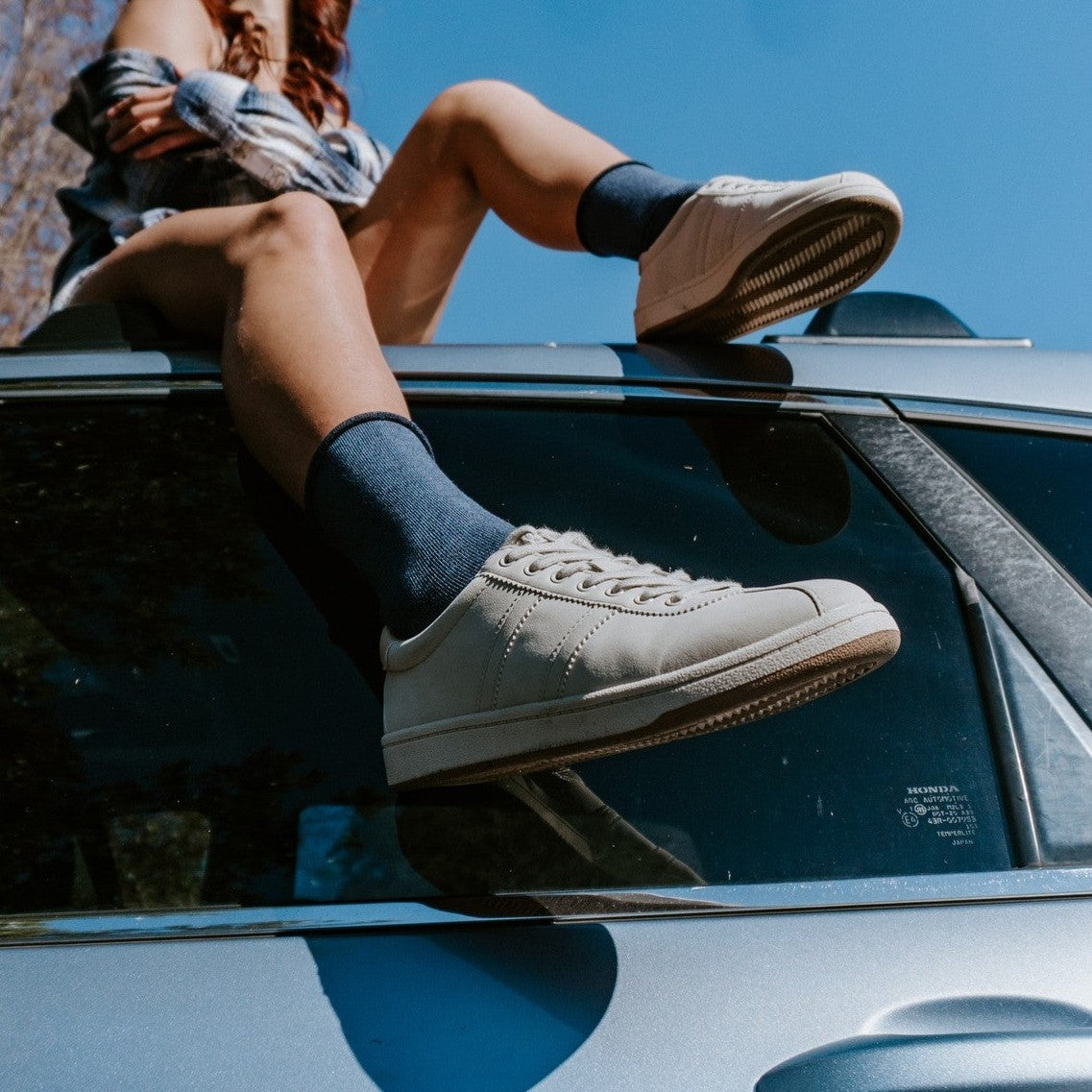 Dramatic low-angle view from beside a Honda car of a woman with crossed legs in navy Solid Bamboo socks and cream lace-up sneakers, reflected in the side window. [lf]