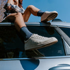 Dramatic low-angle view from beside a Honda car of a woman with crossed legs in navy Solid Bamboo socks and cream lace-up sneakers, reflected in the side window. [lf]