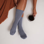 Overhead view of chambray Solid Bamboo socks on a white bed, a rust waffle throw to one side and a glass of coffee held nearby. [lf]