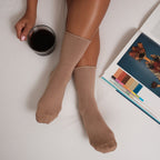 Vertical overhead shot of khaki Solid Bamboo socks on crisp white bedding, the model holding a coffee with a design book open beside her. [lf]