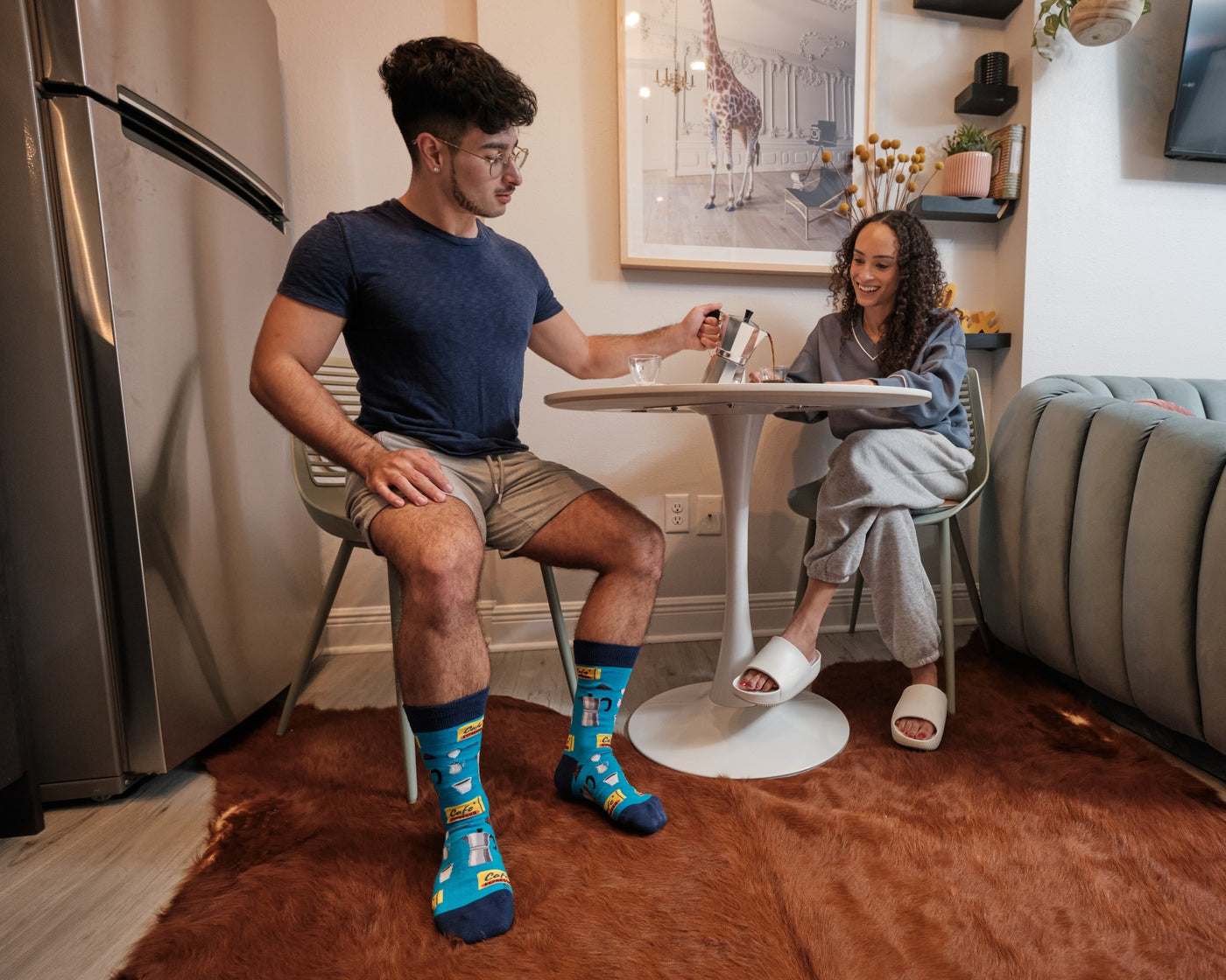 A wide shot of a couple at a small white kitchen table on a brown cowhide rug, the man pouring espresso from a moka pot while wearing Cafe Espresso socks in Blue. [lf]