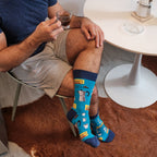A wide cropped shot of socked feet beside a tulip table, Cafe Espresso socks in Blue showing the print clearly next to an espresso glass and white sugar jar. [lf]