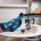 A wide playful shot of feet kicked up onto the table near a stovetop moka pot and a relax sign on the shelf, all framed by the Cafe Espresso socks in Blue. [lf]