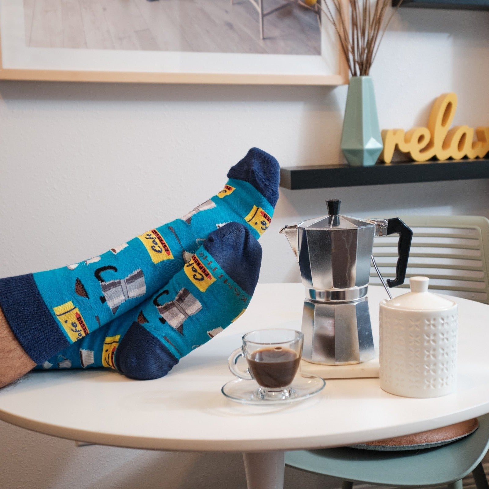 A wide playful shot of feet kicked up onto the table near a stovetop moka pot and a relax sign on the shelf, all framed by the Cafe Espresso socks in Blue. [lf]