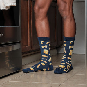 A waist-down kitchen view zooms in on Hasta La Pasta socks in Navy printed with bow-ties, ravioli and farfalle as the wearer stands between dark cabinets and a fridge. [lf]
