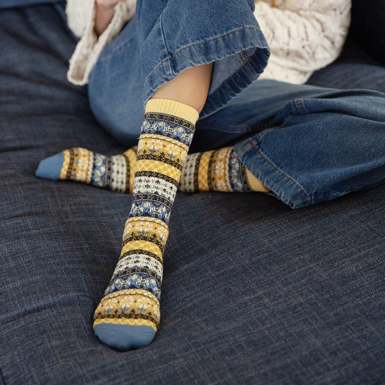 A seated detail of crossed feet in jeans on a navy sofa, focused on the patterned cuff and toe of Honey Bees socks in Yellow against a yellow throw pillow background. [lf]