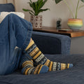 A close in lounging shot of jeans and crossed ankles on a navy chaise beside a wooden side table and potted plants, featuring the bold patterned bands of Honey Bees socks in Yellow. [lf]