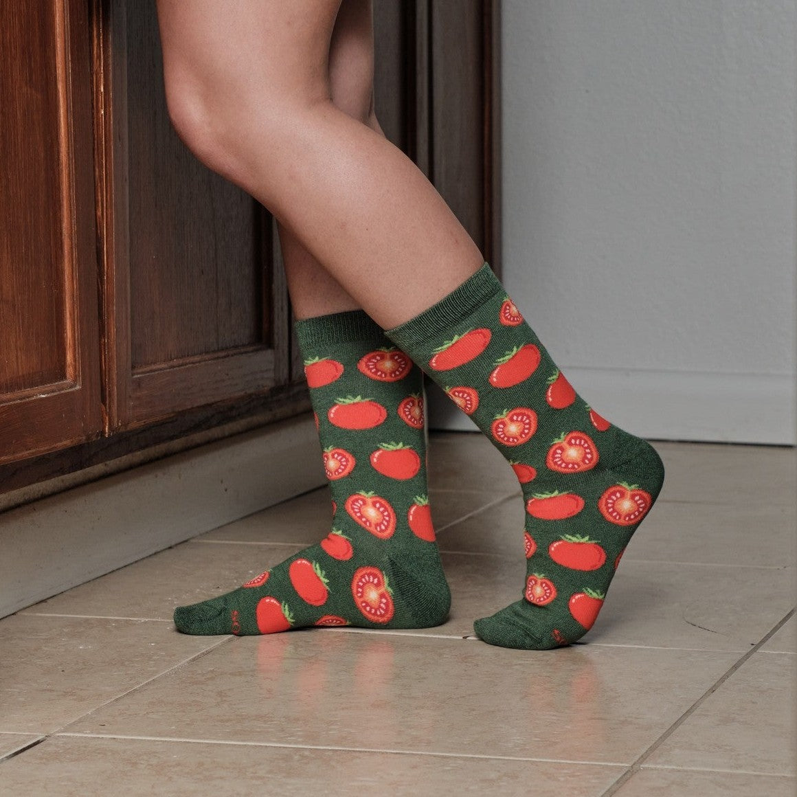 Tight crop on bare legs and ankles posed on kitchen tile by a wood cabinet, putting Tantalizing Tomatoes socks in Black front and center with every tomato print detail in focus. [lf]