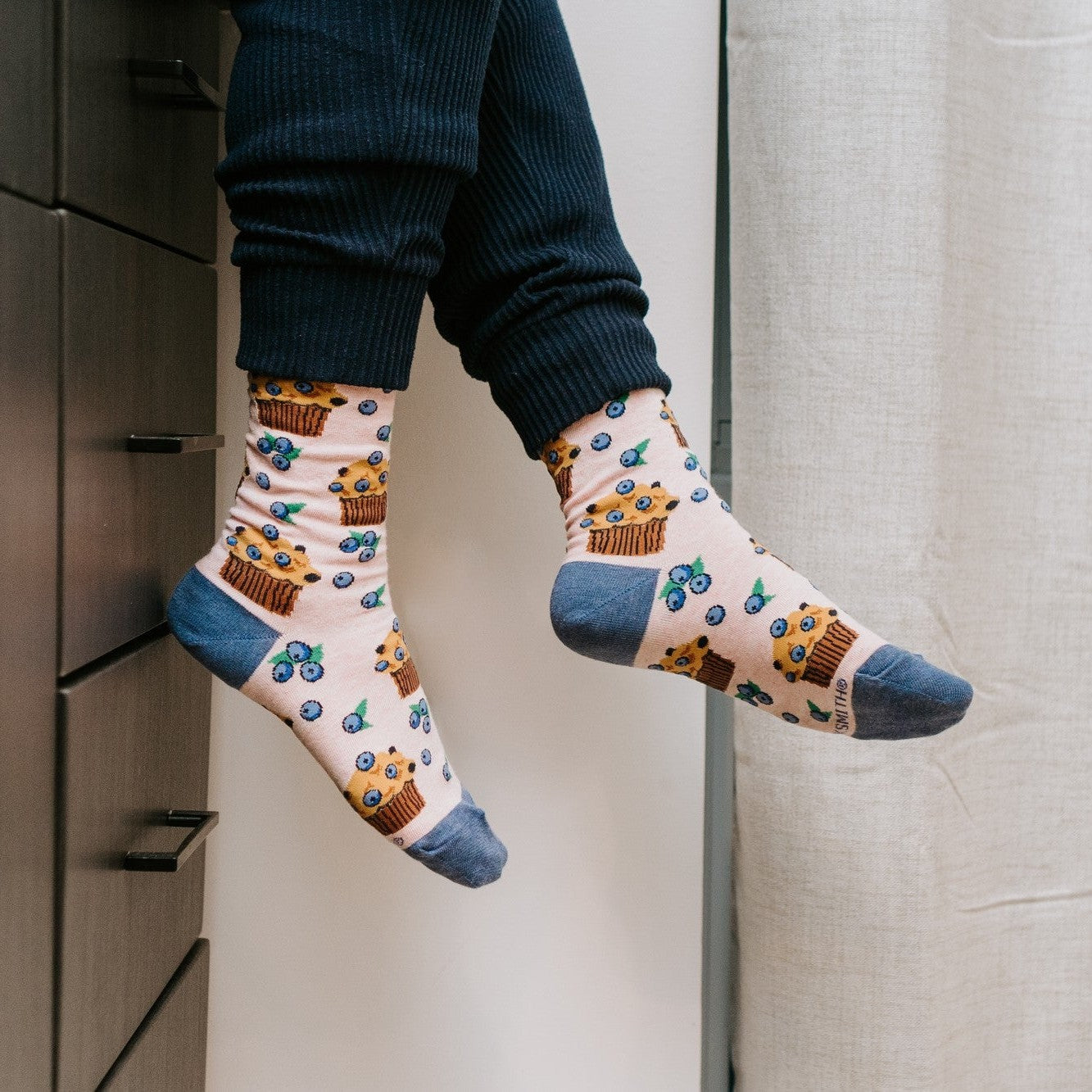 Wide kitchen scene of a smiling model leaning back on the counter with a mug, Berry Tasty Muffins socks in Pink Heather visible at the cuffs of her navy joggers. [lf]