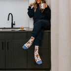 Vertical lifestyle shot of a model perched by the kitchen sink looking up with a mug in hand, Berry Tasty Muffins socks in Pink Heather grounding the cozy navy outfit. [lf]