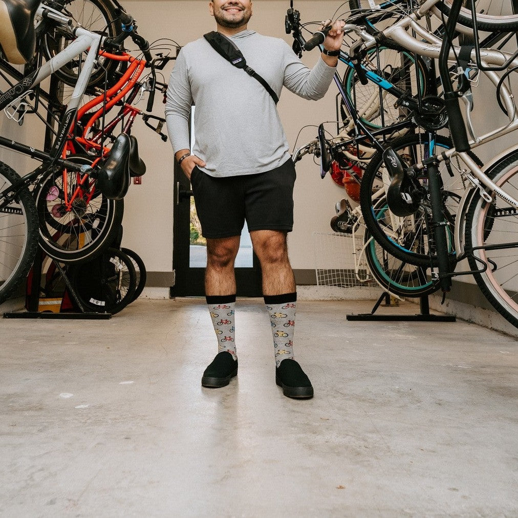 A man stands centered in a corridor of vertically mounted bikes adjusting one with his hand, wearing Bicyle Race Bamboo socks in Light Gray Heather and slip-on sneakers. [lf]