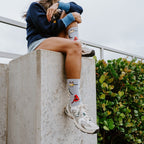 A cropped torso-down shot of a model hugging her knee on a stone ledge, the bird motifs on her Bird Is The Nerd socks in Blue Heather standing out against the soft gray cuff and white trainers. [lf]