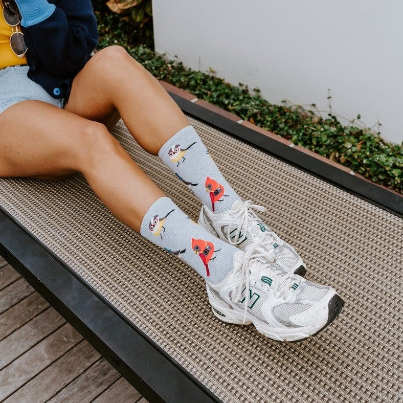A model perched on a chaise lounge points her camera at the viewer, legs stretched out to showcase Bird Is The Nerd socks in Blue Heather paired with white New Balance sneakers and tropical greenery behind her. [lf]