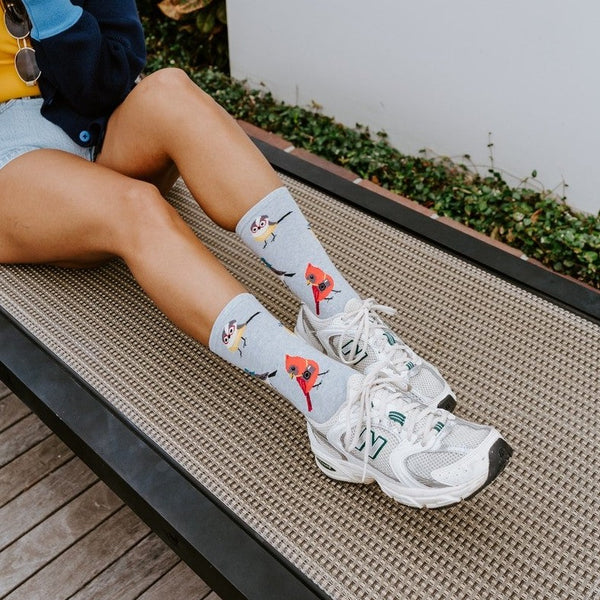 A model perched on a chaise lounge points her camera at the viewer, legs stretched out to showcase Bird Is The Nerd socks in Blue Heather paired with white New Balance sneakers and tropical greenery behind her. [lf]