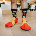 A man in oversized red and yellow polka dot clown shoes stands on a tiled lobby floor showing his Clowning Around Bamboo socks in Black covered in tiny clown faces. [lf]