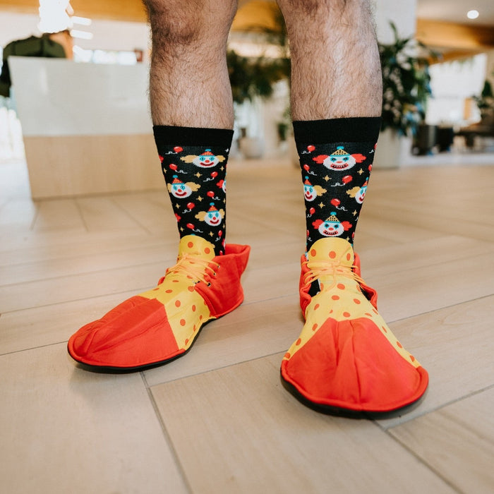 A man in oversized red and yellow polka dot clown shoes stands on a tiled lobby floor showing his Clowning Around Bamboo socks in Black covered in tiny clown faces. [lf]