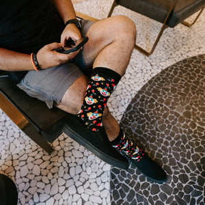 A vertical seated portrait of the same model on his phone on a mosaic floor, crossed legs spotlighting his Clowning Around Bamboo socks in Black against black sneakers. [lf]