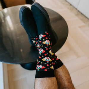 A relaxed vertical frame from beside the round side table showing the model's Clowning Around Bamboo socks in Black with a leather lounge chair and orange cushion in view. [lf]