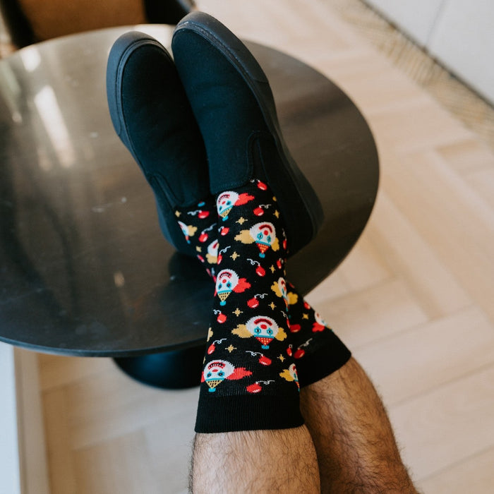 A relaxed vertical frame from beside the round side table showing the model's Clowning Around Bamboo socks in Black with a leather lounge chair and orange cushion in view. [lf]