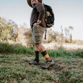 Smiling man in a straw hat and brown long-sleeve top hikes across a grassy field with a loaded backpack, his Grand Canyon National Park socks in Green Heather visible above hiking boots. [lf]