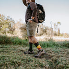 Smiling man in a straw hat and brown long-sleeve top hikes across a grassy field with a loaded backpack, his Grand Canyon National Park socks in Green Heather visible above hiking boots. [lf]