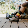 Casual deck scene of a man seated with knees bent and arms resting, the Grand Canyon National Park socks in Green Heather showing the colorful park graphic. [lf]