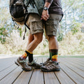 Two hikers stand side by side on a deck holding hands, both wearing Grand Canyon National Park socks in Green Heather with their hiking boots. [lf]