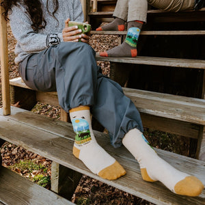 A woman in a cozy sweater sips a hot drink on cabin steps, her Yellowstone National Park socks in Yellow paired with her partners Brown Heather above. [lf]
