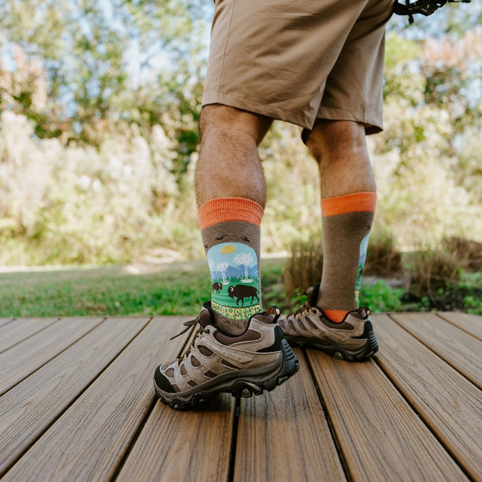 Low-angle calf shot of a man in tan shorts and Merrell hikers, Yellowstone National Park socks in Brown Heather showing the park scene cuff. [lf]