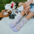 Tall vertical shot of legs stretched out in Honey Bee Floral socks in Blue Heather, with a pink and white bouquet, polaroid photos, and an instant camera resting on a textured blanket. [lf]