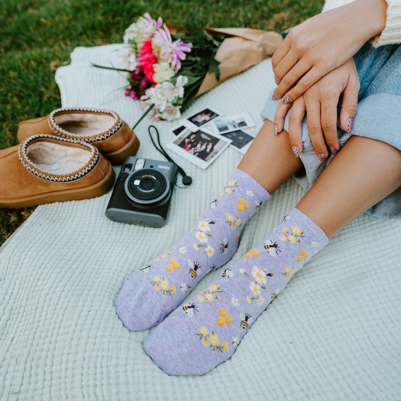 Tall vertical shot of legs stretched out in Honey Bee Floral socks in Blue Heather, with a pink and white bouquet, polaroid photos, and an instant camera resting on a textured blanket. [lf]