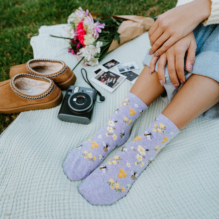 Tall vertical shot of legs stretched out in Honey Bee Floral socks in Blue Heather, with a pink and white bouquet, polaroid photos, and an instant camera resting on a textured blanket. [lf]