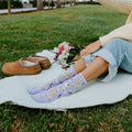 Woman in cream sweater raises an instant camera overhead for a selfie while sitting on a blanket in a green park, wearing Honey Bee Floral socks in Blue Heather. [lf]