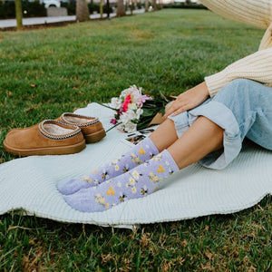 Woman in cream sweater raises an instant camera overhead for a selfie while sitting on a blanket in a green park, wearing Honey Bee Floral socks in Blue Heather. [lf]
