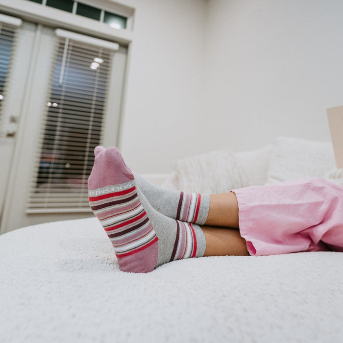 A low side-on shot from the cushion catches a reader propping up her feet, the soles of her Sport Stripe socks in Light Gray Heather facing the camera with the Socksmith logo visible. [lf]
