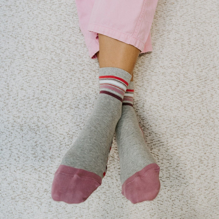 A top-down view of relaxed pink-clad legs on a boucle blanket centers Sport Stripe socks in Light Gray Heather with their layered stripe band and contrast pink toe. [lf]