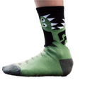 Lifestyle shot of a child wearing a pair of green and black T-Rex socks on a tree stump. [lf]