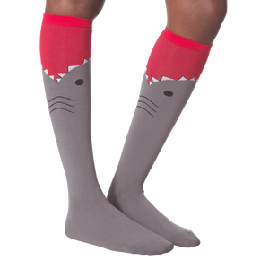 A pair of gray knee-high socks with a red cuff. The socks have a cartoon shark face on them, with the shark's mouth open and its teeth showing.