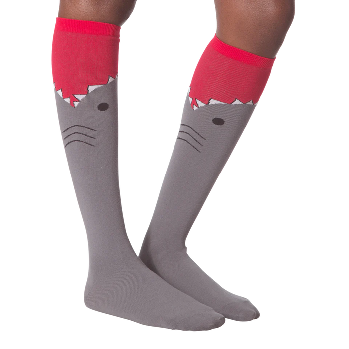 A pair of gray knee-high socks with a red cuff. The socks have a cartoon shark face on them, with the shark's mouth open and its teeth showing.